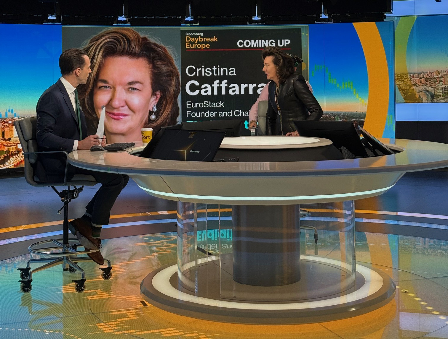 TV studio with host on one side of table and Cristina Caffarra on the other. In the background is a large screen which reads 'Bloomberg Daybreak Europe – Coming up – Cristina Caffarra