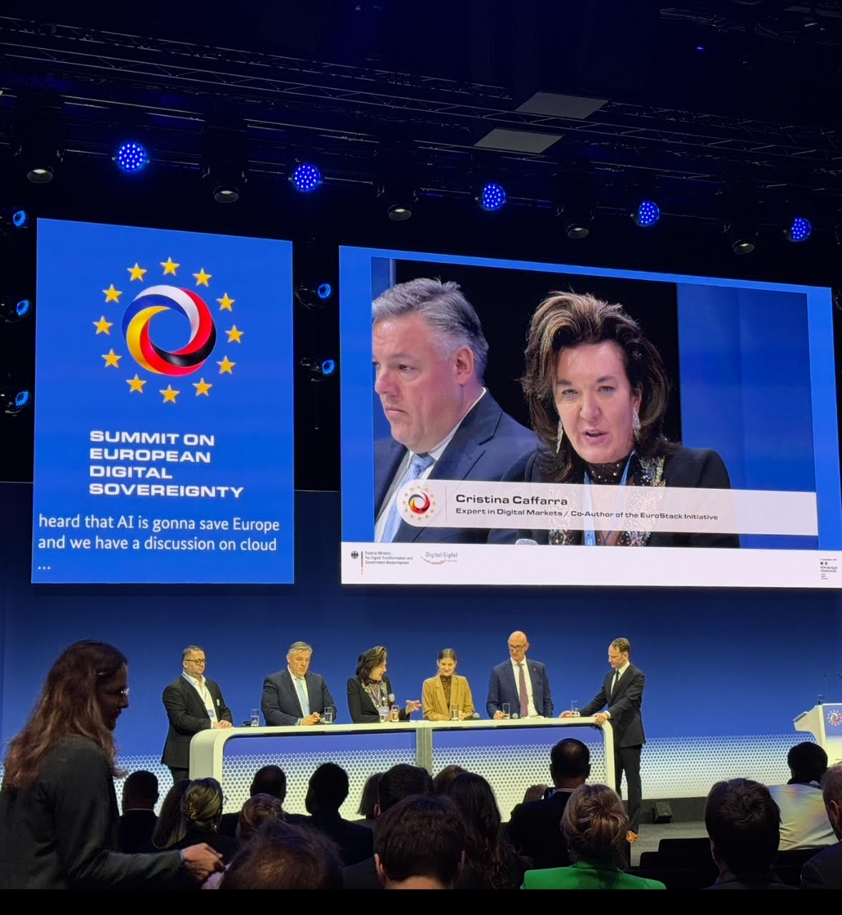 Panel of speakers in front of screen reading 'Summit on European Digital Sovereignty' with large image of Cristina Caffarra projected on the screen